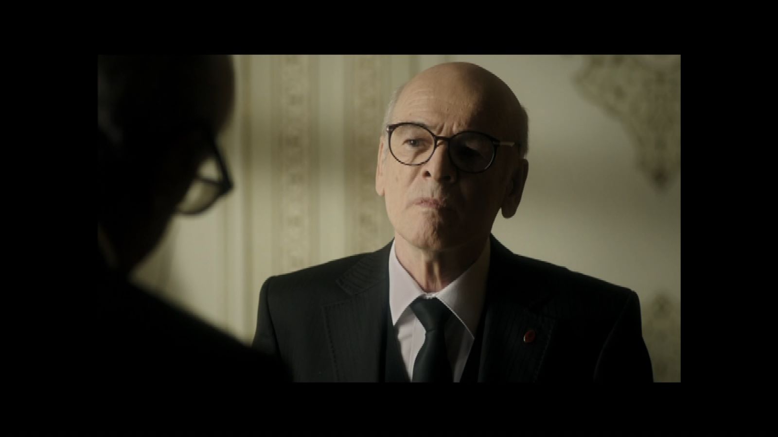 Ben Onu Çok Sevdim — A bald man in glasses and a suit facing another man indoors.