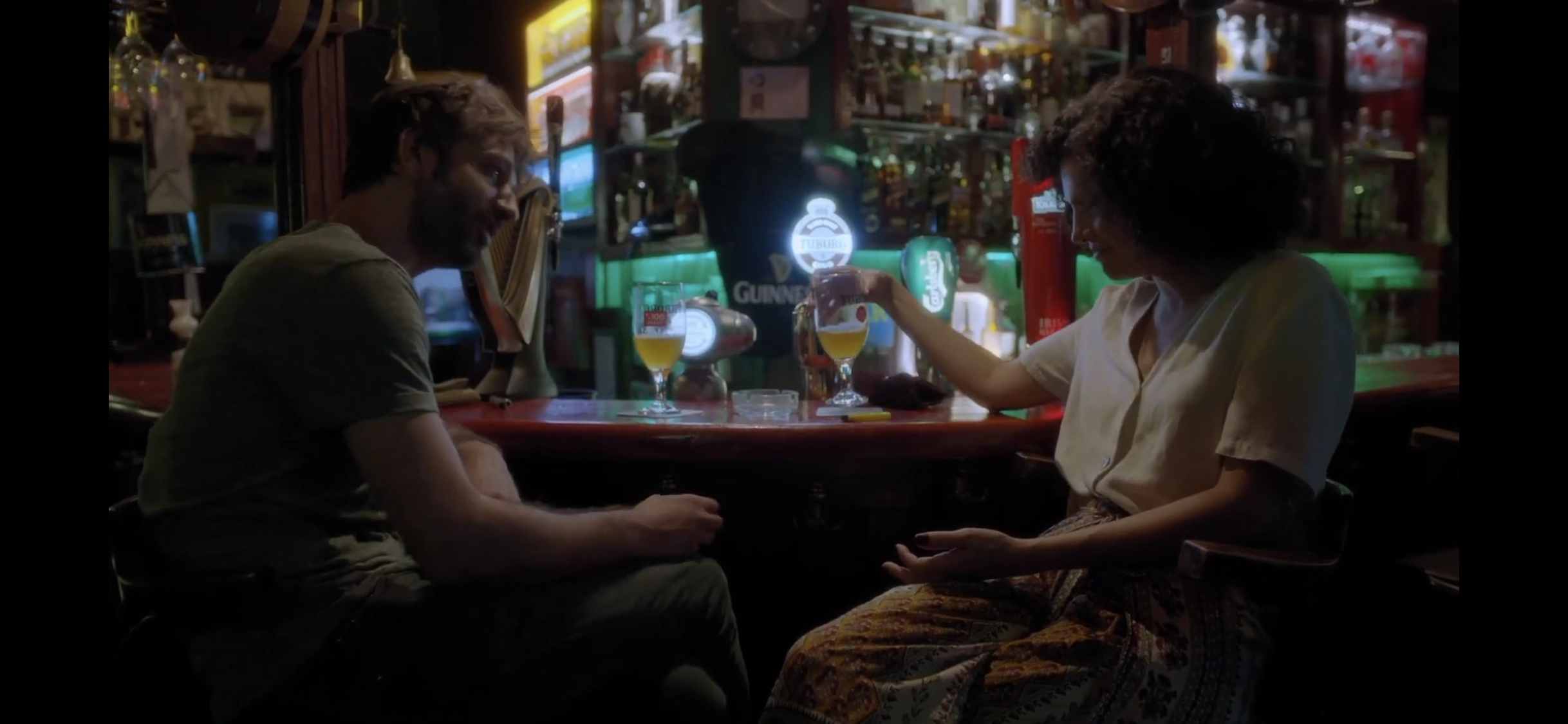 Diyalog — A man and a woman talking across a bar counter beneath colourful lights.
