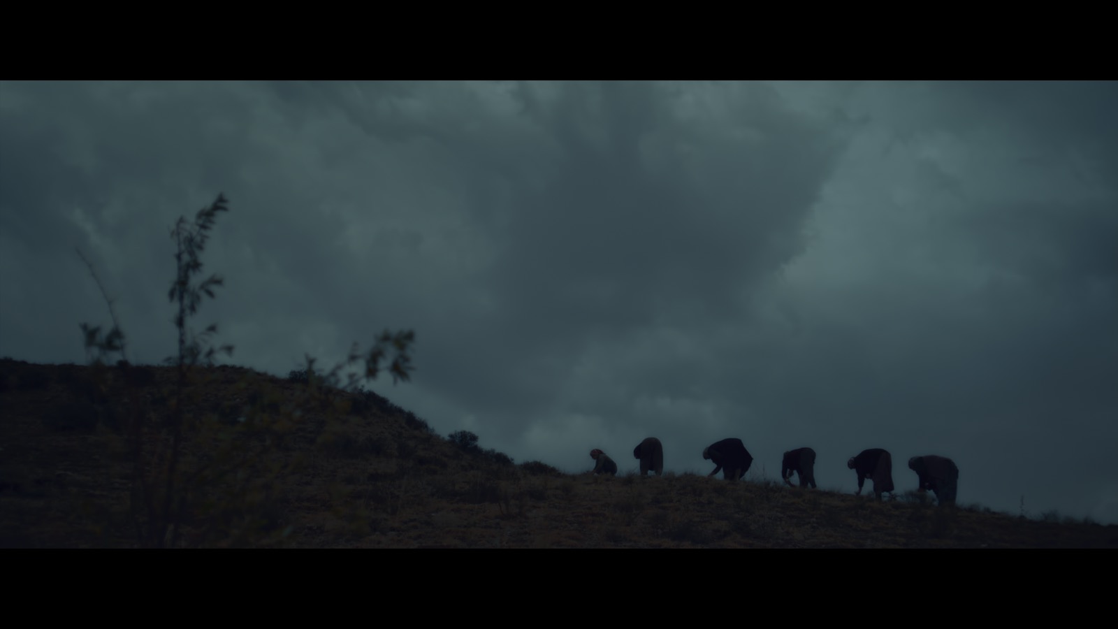 Kabahat — Figures working on a hillside beneath a dark, cloudy sky.