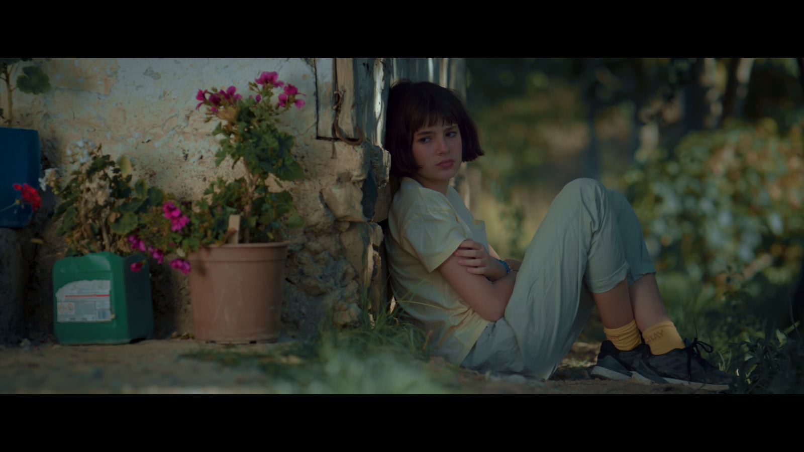 Kabahat — A girl sitting against a stone wall beside flowering pots.