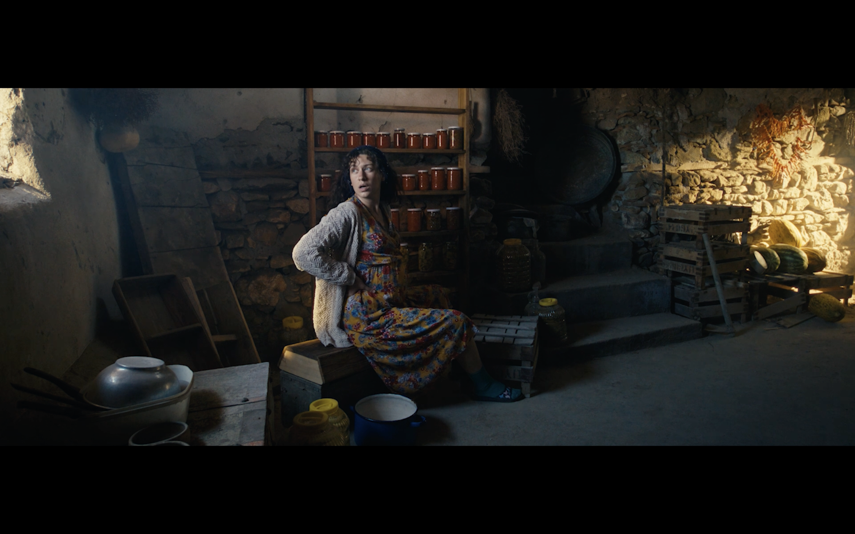 Kavanoz — A woman seated in a stone-walled room with jars on a shelf.