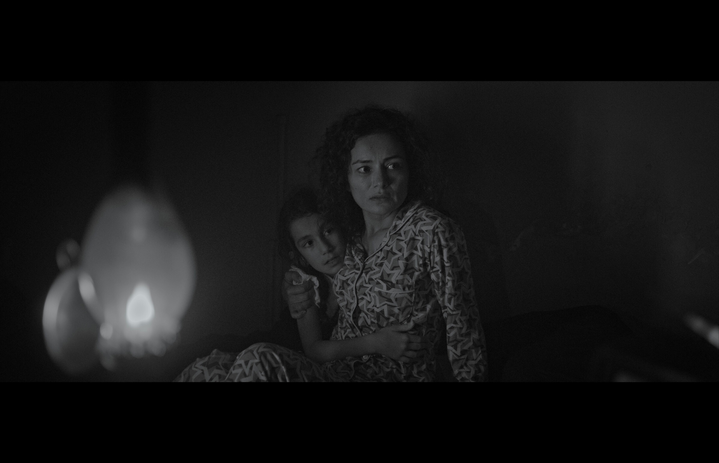 Sığınak — Black-and-white frame. A woman seated beside the glow of an oil lamp.