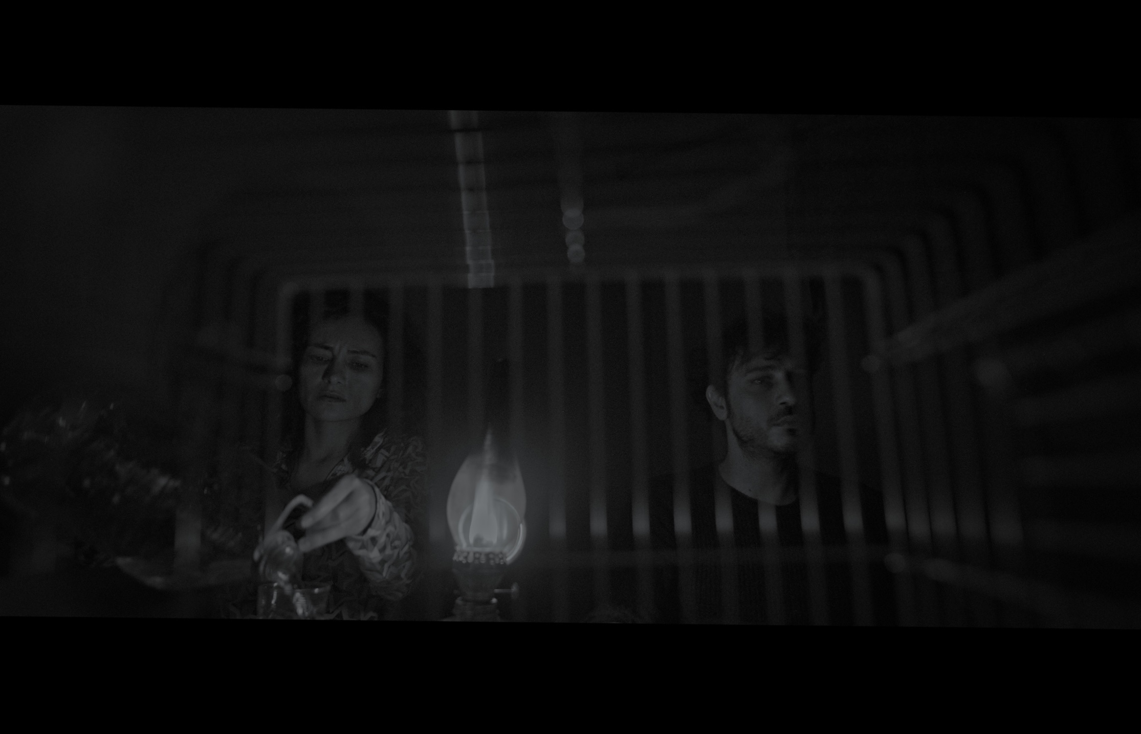 Sığınak — Black-and-white frame. Two figures and a lamp seen through a striped translucent foreground.